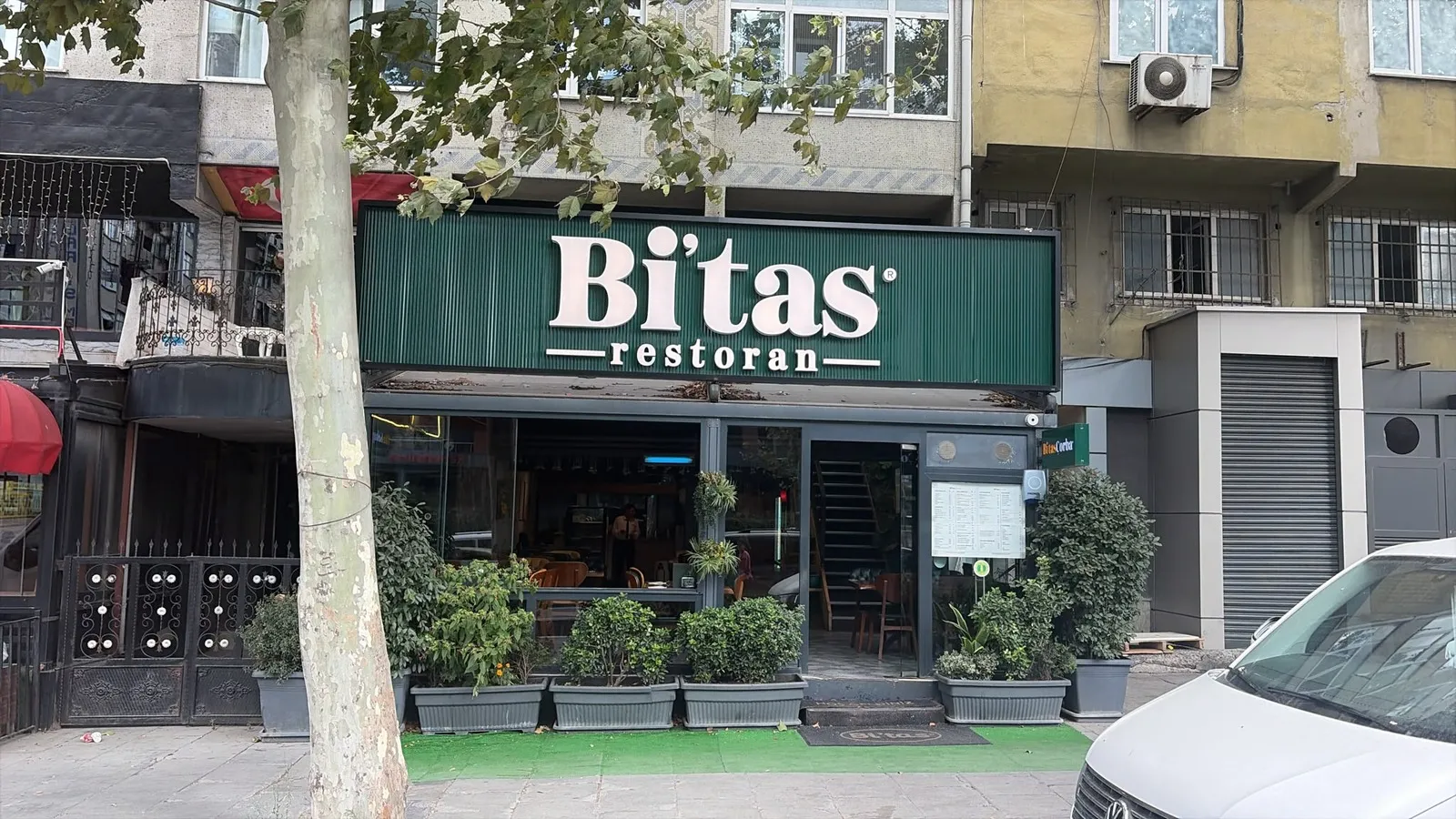 Bi'tas Restoran'ın cadde üzerindeki dış cephesi; yeşil tabelada "Bi'tas restoran" yazıyor, girişte saksı bitkiler